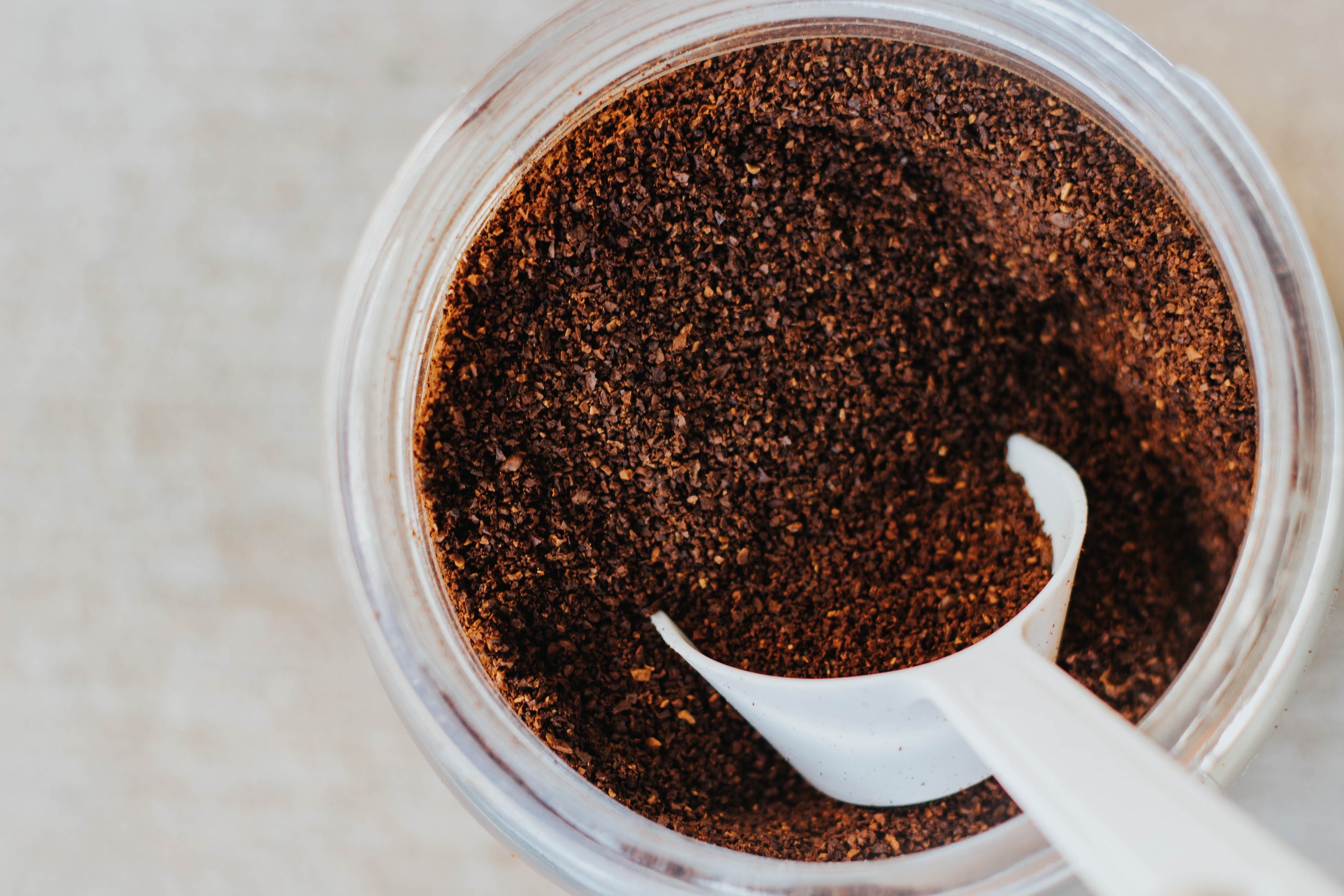 This is a top-down, close-up shot of a clear glass jar filled with dark brown, coarsely ground coffee. The jar appears to be almost entirely full, with the coffee granules creating a textured, uneven surface. The coffee itself has a rich, chocolatey hue with subtle variations in shade, suggesting a blend of different beans or roast levels. A white plastic scoop is partially submerged within the coffee grounds, its curved shape contrasting with the granular texture of the coffee. The scoop is positioned towards the lower right of the frame, appearing to have just scooped up a portion of the coffee. The jar’s glass is clean and transparent, allowing a clear view of the coffee within. The background is a light beige or off-white surface, possibly a countertop or table, providing a neutral backdrop that emphasizes the coffee and jar. The lighting is soft and diffused, creating gentle shadows and highlighting the texture of the coffee grounds. The overall composition is simple and focuses on the raw, natural element of coffee, evoking a sense of warmth and aroma. The image suggests a moment of preparation or enjoyment related to coffee brewing.